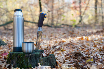 Obraz premium Thermos and hunting knife. The concept of tourism, survival and hunting in the autumn in forest. a thermos of tea, a cup and a knife, on an old tree stump. dry leaves in the park. at nature. close-up