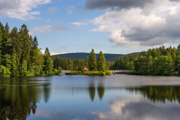 Obraz premium Picturesque view of small lake in forest. Wooden bridge, gazebo for rest. Blue sky and trees reflected in water. High quality photo.