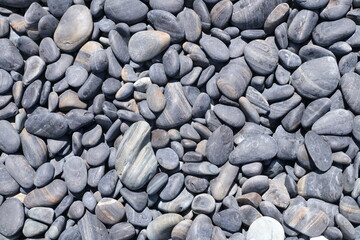 A detailed view of a bed of beach stones. The stones are various sizes and shapes, with a predominantly dark gray hue. The image highlights the natural textures and patterns formed by years of weather