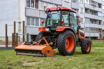 Large orange tractor moving green farmers pasture. Tractor with a grass mower in the city. Mowing the silage