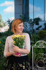 Beautiful middle-aged woman holds a pot with yellow chrysanthemum flower front of the modern building with flower shop. Selective focus.