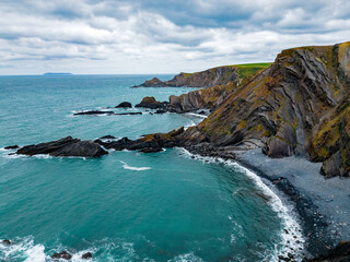 Scenic North Devon coastline with shingle beach and beautiful rock formations. Aerial view at Eye Cove on a cloudy day. Rocks covered with moss. Lundy island at distance. Ocean waves. © Ssisabal