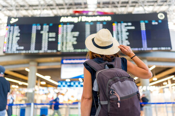 A woman wearing a straw hat and a backpack is looking at a large airport screen