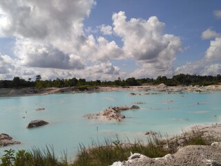 A picturesque view of a turquoise lake within a limestone quarry, surrounded by rugged terrain and lush greenery. The vibrant blue water contrasts beautifully with the dramatic cloud-filled sky.