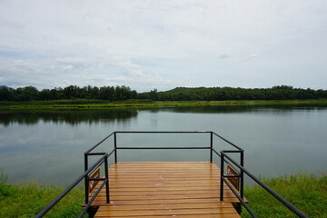 A tranquil scene of a wooden pier extending into a calm lake. The pier has a metal railing and offers a peaceful view of the surrounding natural scenery. Lush green hills and a cloudy sky complete the