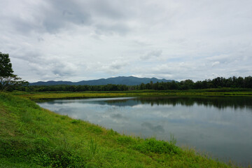 A serene landscape photo showcasing a lake surrounded by lush greenery and mountains in the distance. The sky is overcast with soft, white clouds. The lake is calm and reflective, mirroring the beauty