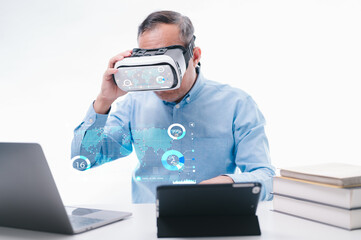 A man wearing a VR headset is sitting at a desk with a laptop and tablet. There are several books stacked on the desk, and a keyboard is visible