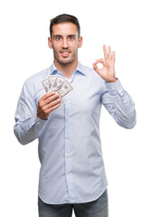 Handsome young man holding money doing ok sign with fingers, excellent symbol