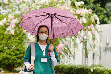 Nurse wearing medical mask holding pink umbrella, standing in hospital garden with blooming flowers behind. She has name badge around neck and looks professional and composed