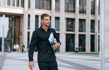 young man in a black shirt walks confidently while holding documents and a smartphone, embodying a successful and focused professional on the move in an urban setting