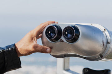 Close up of a hand adjusting silver binoculars against a clear blue sky. The binoculars are mounted an observation point for tourists to view distant landscapes. Man uses a coin operated telescope