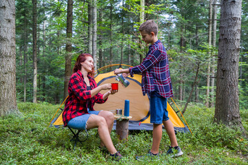 Fototapeta premium A family is relaxing at a campsite in the forest. Mother and son drink coffee near the tent.