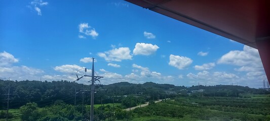 Bright blue sky with fluffy white clouds over a peaceful rural landscape. Lush green hills, a winding road, and power lines define the tranquil countryside