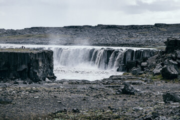 Waterfall cascading over rugged basalt cliffs in an expansive volcanic landscape under a cloudy sky, capturing the raw power and isolation of Icelandic nature.