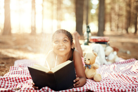 Happy girl, book and forest with light for fairytale, picnic or magical moment in nature. Young female person, little child or kid with smile on blanket for fairy story, sparkle or fantasy in woods
