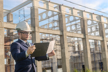 Asian adult foreman stands in front of the construction inspecting a construction project with...