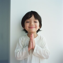 A young child with clasped hands and an endearing smile, dressed in white, radiates purity and contentment against a simple, soft background.