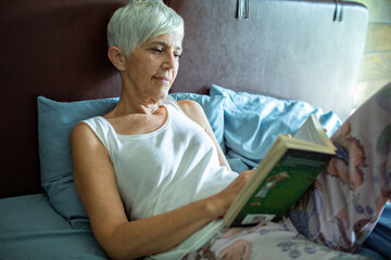 Senior woman laying in bed reading a book at home