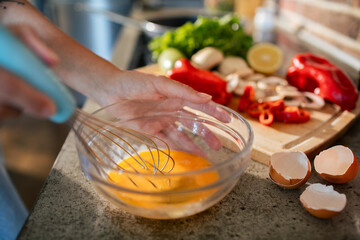 Close up of woman whisking eggs with fresh vegetables in the background in kitchen
