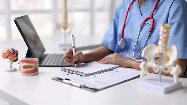 Medical professional taking notes at desk with anatomical models. Doctor in a medical office writes notes on a clipboard, surrounded by anatomical models of a brain, teeth, and spine.