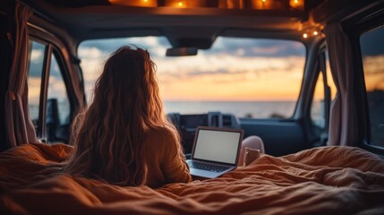 Fototapeta premium A woman relaxes in a warm camper van, illuminated by string lights, as she works on her laptop while watching a breathtaking sunset