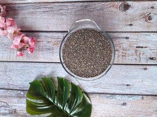 Glossy Chia Seeds in a Glass Bowl on Wooden Surface, Accompanied by Scattered Seeds and a Decorative Touch of Pink Flowers in the Background.