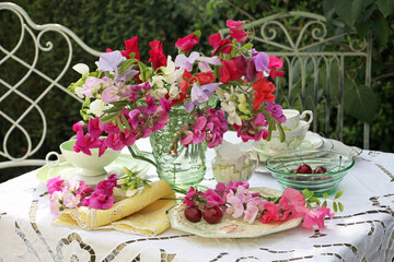 Sweet peas in a glass jug floral still life on a table set for afternoon tea outdoors in a garden