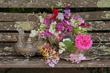 still life of roses and sweet peas and honeysuckle in antique silver vases on a rustic wooden bench outdoors