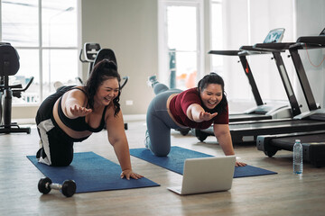 Two women exercising together on yoga mats in a gym, following an online workout on a laptop, surrounded by gym equipment.