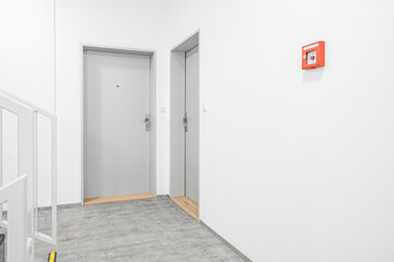 A well-lit hallway with closed gray doors and a red emergency fire alarm box on a white wall,...
