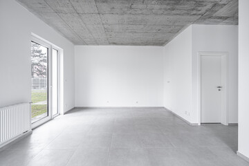 Empty modern room with large glass door, concrete ceiling, and neutral tile flooring, featuring...