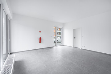 Bright, empty room with gray tiled floor, white walls, and large windows, featuring a door leading outside. A fire extinguisher is mounted on the wall, adding a safety element to the modern space.