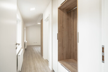 A bright, minimalistic hallway in an apartment with built-in wooden cabinets, light-colored walls, and windows, leading to another room.