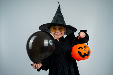 Portrait of a little Caucasian girl in a witch costume holding a cauldron and a black balloon on a white background. 