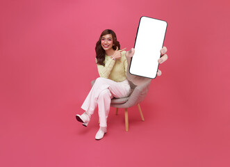 Full length beautiful Asian teen woman using smartphone and sitting on chair isolated on pink copy...