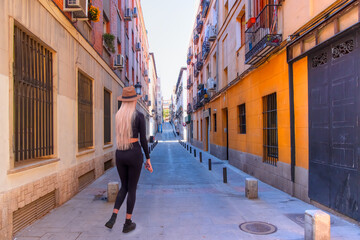 A blonde woman wearing a stylish hat walking narrow street in old Madrid downtown with colorful typical houses - Madrid, Spain