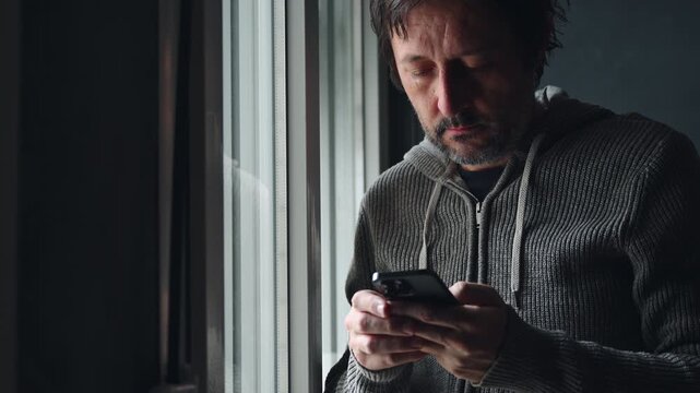 Man using mobile smartphone for texting messages, adult male person standing by the window of dark living room sending SMS with smart phone app,