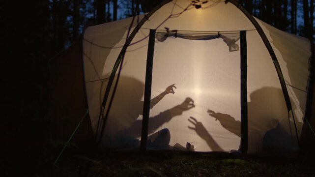 Silhouettes of children playing in camping tent at night making shadow puppets with flashlight during summer holidays
