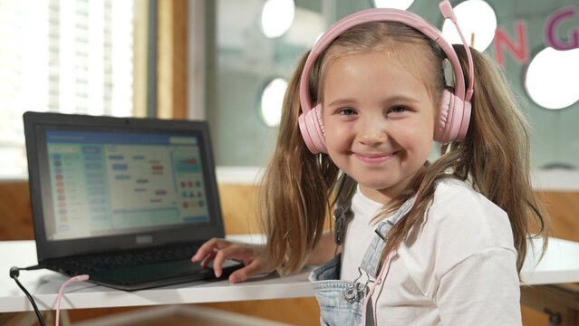 Smiling smart caucasian girl show system programing or coding program in STEM technology education. Child wearing headphone while working by using laptop writing code and look at camera. Erudition.