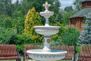 White three-tiered fountain on church grounds. Painted patterns on falling water object in public park. Stone material and design fountain details on built structure. Famous place of leisure activity.