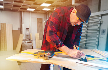 Man carpenter at work. Guy who makes furniture is standing in his workshop. Carpenter makes marks on boards. Man furniture maker in checkered shirt. Carpenter working in production workshop