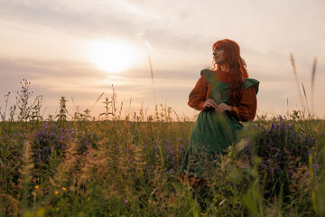 A shepherdess woman with red hair and a red and green dress walks on a field of flowers. Rural...