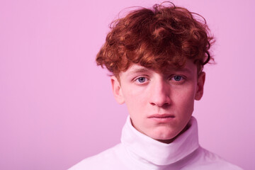 Portrait of young man with curly red hair standing against pink background wearing white turtleneck with thoughtful expression looking straight at camera