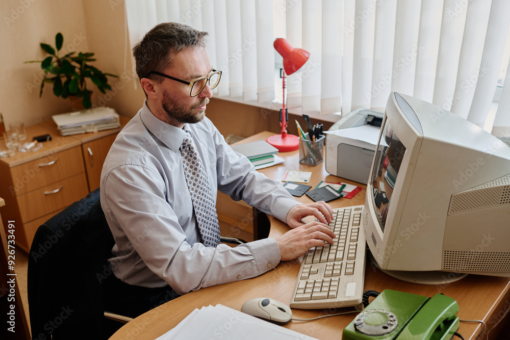 Poster Caucasian man wearing glasses working at desk in office environment with desktop computer and various office supplies on desk surrounded by files and paperwork