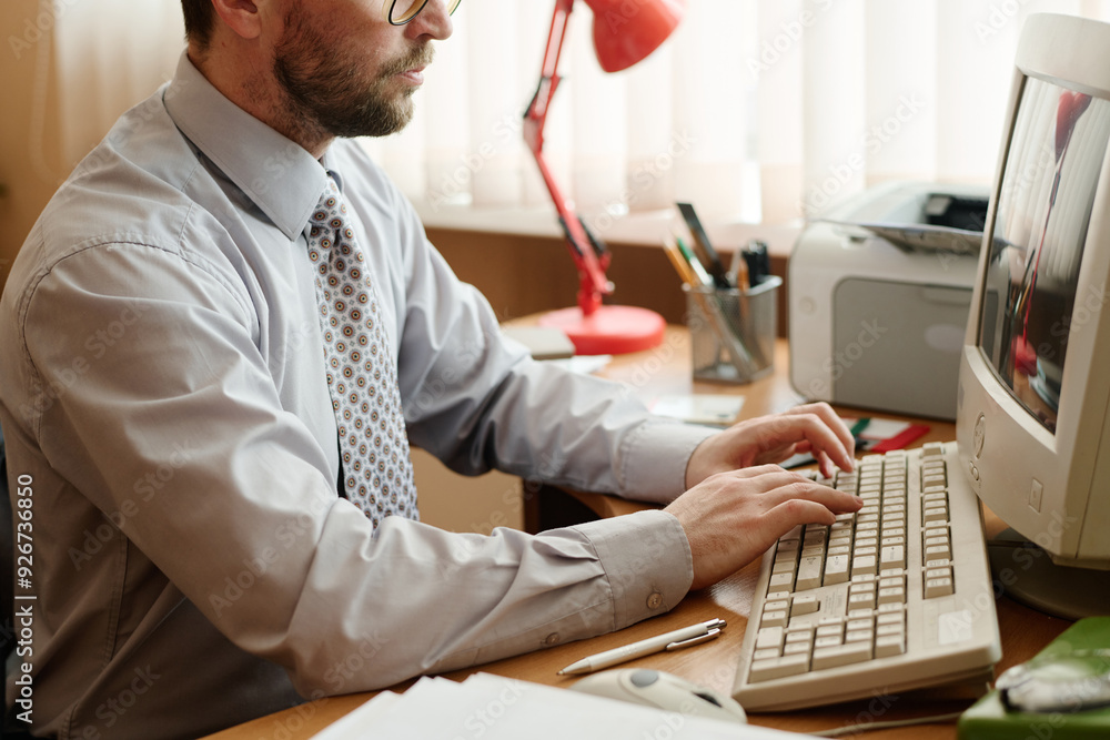 Poster man sitting at desk, using computer while writing, wearing glasses and formal attire. lamp, stapler,