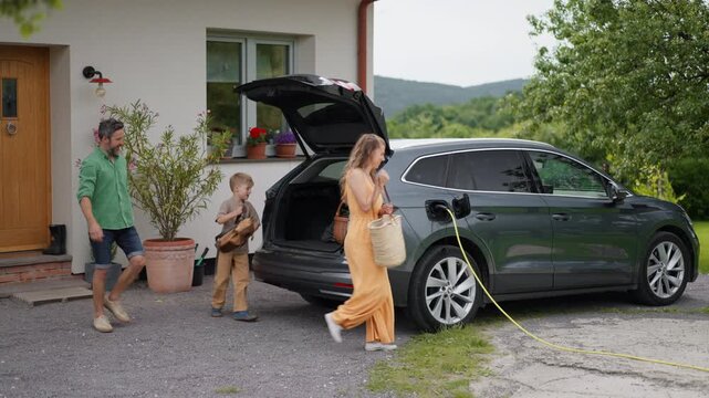 Family loading luggage into trunk of electric car with charger in charging port. Travelling on family road trip or vacation by electric vehicle.