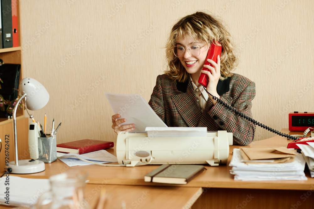 Poster Caucasian female smiling while using retro red rotary phone at desk with office supplies and documents. Dressed in business attire and appearing happy in professional work environment