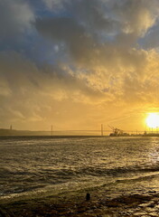 Sunset over Lisbon's 25 de Abril Bridge with dramatic clouds and a river reflecting the golden light