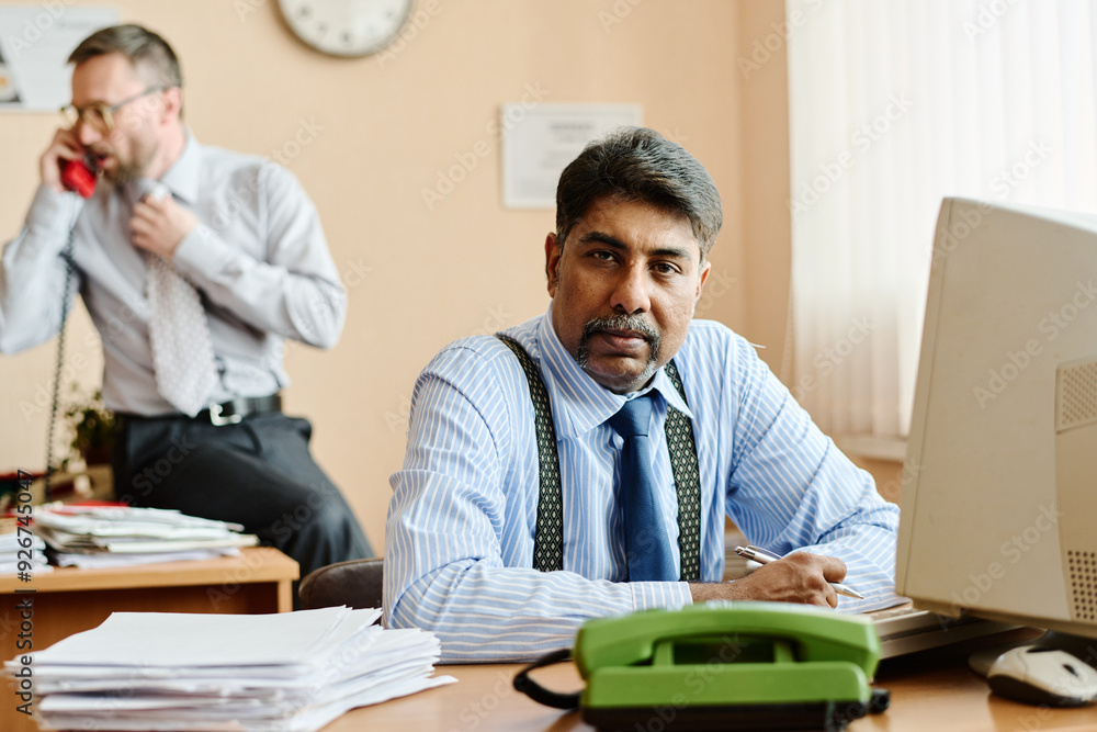 Poster Portrait of businessman sitting at desk, focusing on work while colleague talks on phone in background, office environment with various documents and devices present