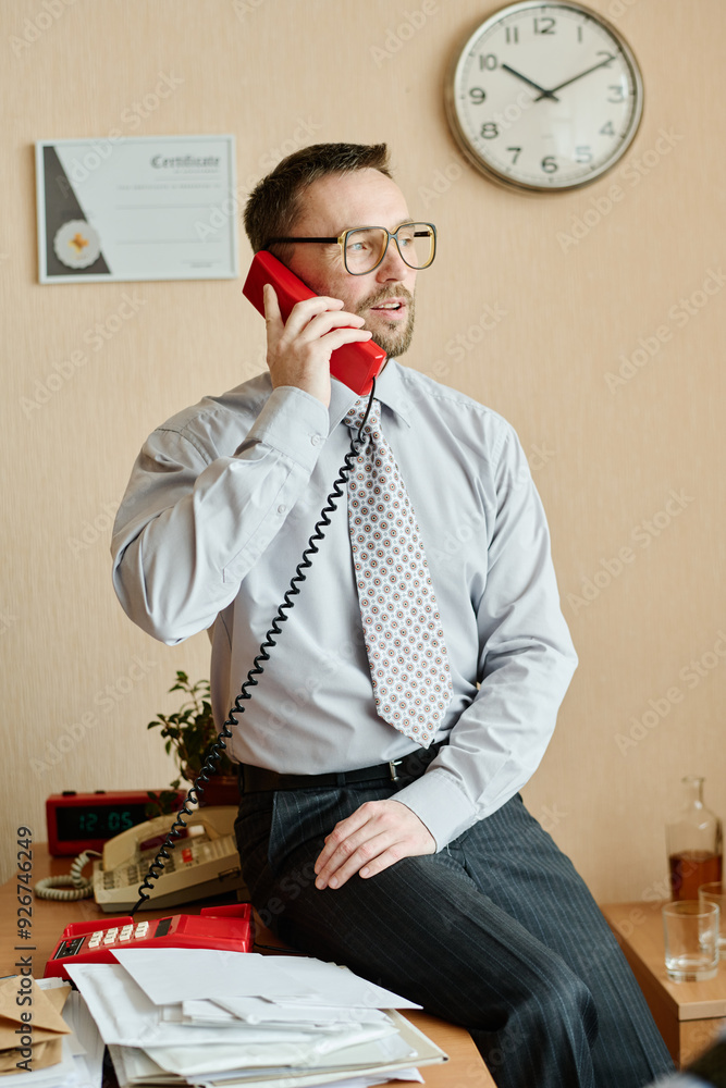 Poster man in business attire holding telephone, conversing while sitting at desk with documents. several o